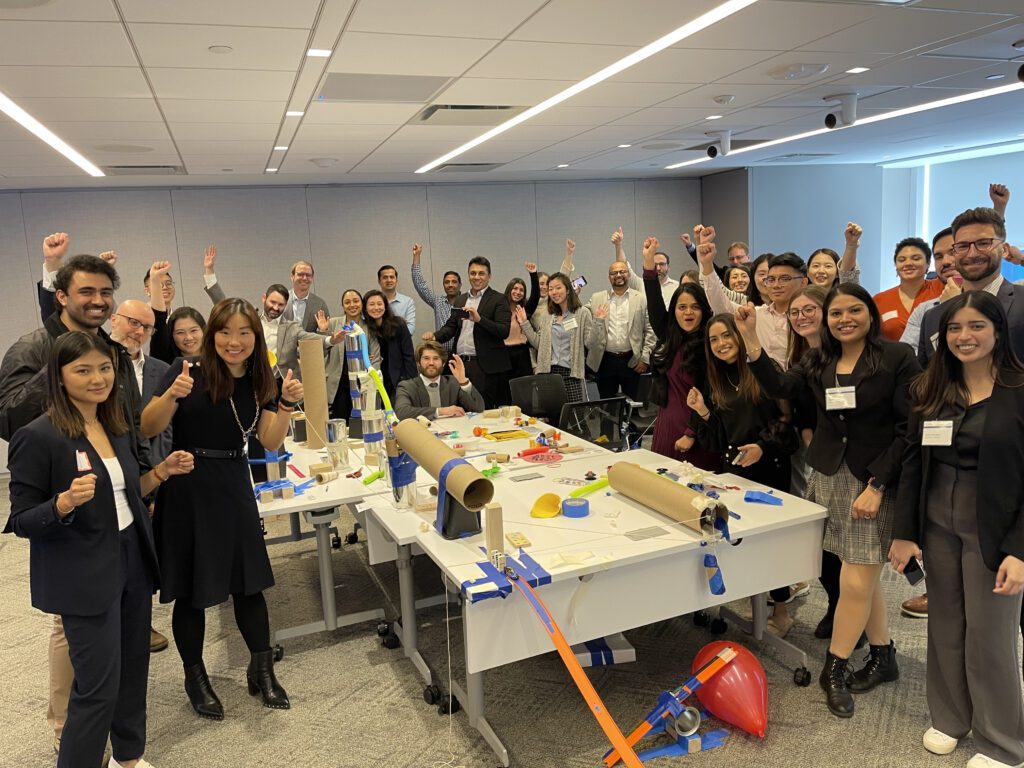 Group of participants celebrate the completion of their collaborative Chain Reaction machine during a team building event, gathered around a complex setup of interconnected parts on a table.