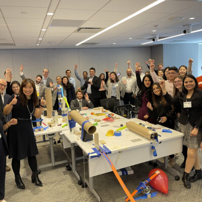 Group of participants celebrate the completion of their collaborative Chain Reaction machine during a team building event, gathered around a complex setup of interconnected parts on a table.