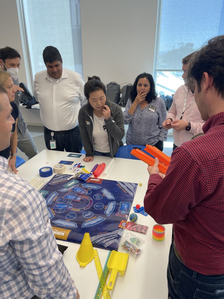 A focused team collaborates around a table filled with materials and a game board for a chain reaction team building activity, strategizing on how to create an interconnected setup to achieve a common goal.