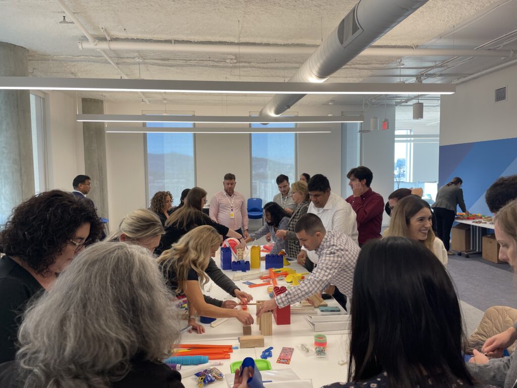 Participants in a team building activity collaborate to create a chain reaction setup. The group is fully engaged, working together with various materials on a shared table, fostering teamwork and creativity.