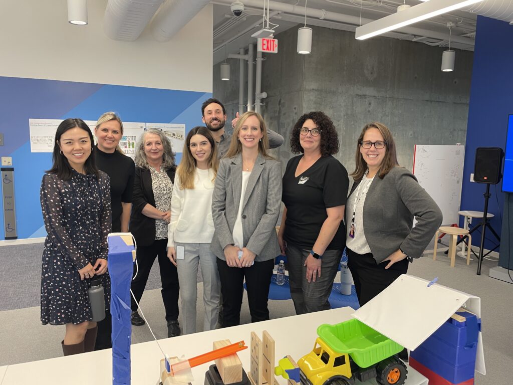 Group photo of team members standing together behind a chain reaction setup during a team building event. The participants are smiling, showcasing a collaborative and friendly atmosphere.