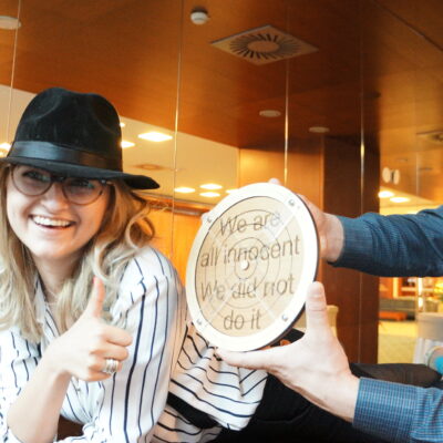 Participants in themed costumes, including hats and suspenders, holding up a clue that reads 'We are all innocent, We did not do it' during the 'Escape the Mob' team building event.