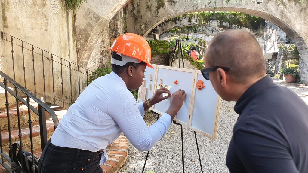 Outdoor team building exercise focused on communication and problem-solving. A participant wearing a hard hat carefully assembles a model while receiving guidance from a teammate, emphasizing teamwork and strategy.