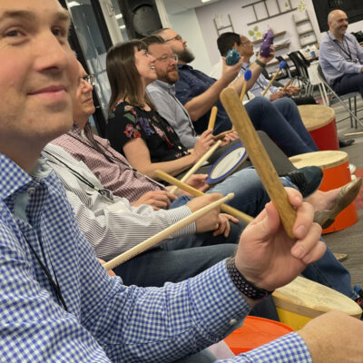 Participants seated in a circle, practicing rhythm on percussion instruments during a BeatsWork team building activity.