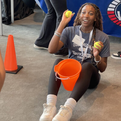 A participant sitting on the floor holding two tennis balls and an orange bucket, engaging in a challenge during the In It to Win It team building event, demonstrating focus, coordination, and a playful spirit.