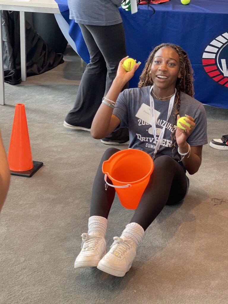 A participant sitting on the floor holding two tennis balls and an orange bucket, engaging in a challenge during the In It to Win It team building event, demonstrating focus, coordination, and a playful spirit.