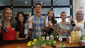 A group of participants smiling and holding various bottles and cocktail-making tools during the Team Mixology team building activity, surrounded by ingredients and mixers, ready to learn the art of crafting cocktails.