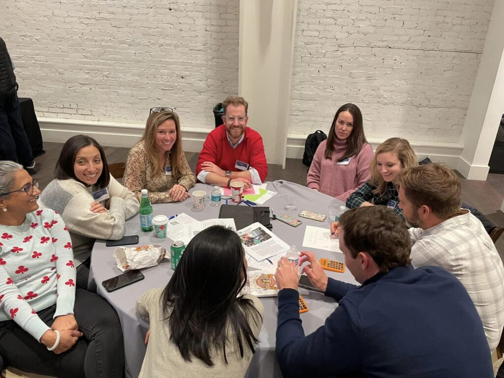 A group of participants sits around a table, smiling and engaged in conversation during the Helping Hands team building event. They are working together on tasks related to assembling care kits for the community.