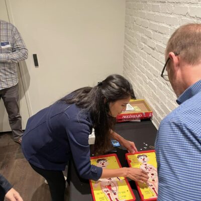 Participants engage in a fun challenge, playing the game 'Operation' as part of the Helping Hands team building event. The activity is designed to earn supplies for care kits through interactive and skill-based tasks.