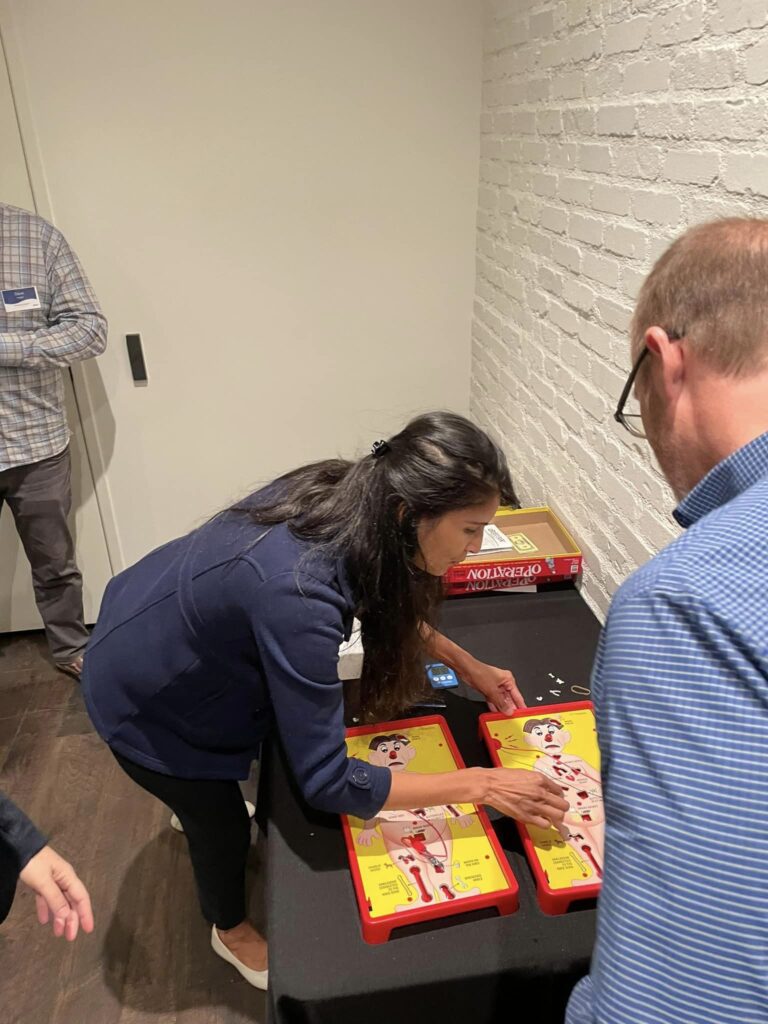 Participants engage in a fun challenge, playing the game 'Operation' as part of the Helping Hands team building event. The activity is designed to earn supplies for care kits through interactive and skill-based tasks.