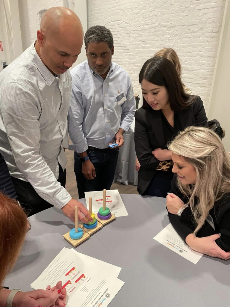 Participants gather around a table, engaged in a problem-solving activity involving colorful rings and pegs during the Helping Hands team building event. The group is focused and collaborating to complete the challenge.