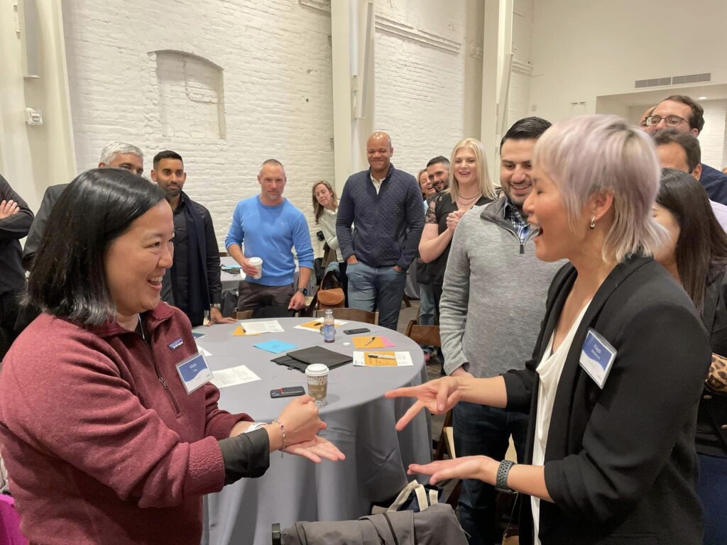 Two pTwo participants enthusiastically engage in a rock-paper-scissors game while others watch and smile during the Helping Hands team building event. The room is filled with energy as the group enjoys the interactive activity.articipants enthusiastically engage in a rock-paper-scissors game while others watch and smile during the Helping Hands team building event. The room is filled with energy as the group enjoys the interactive activity.