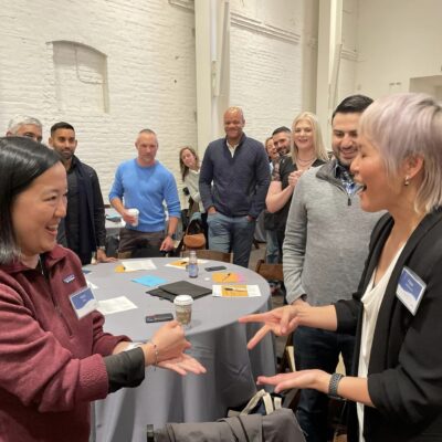 Two pTwo participants enthusiastically engage in a rock-paper-scissors game while others watch and smile during the Helping Hands team building event. The room is filled with energy as the group enjoys the interactive activity.articipants enthusiastically engage in a rock-paper-scissors game while others watch and smile during the Helping Hands team building event. The room is filled with energy as the group enjoys the interactive activity.