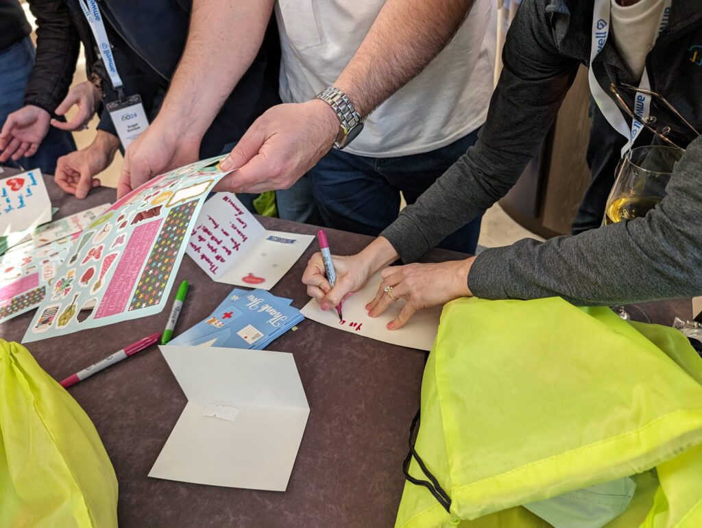 Participants are seen decorating cards with stickers and writing messages for healthcare professionals as part of the Healthcare Heroes scavenger hunt team building event. Bright green donation bags are on the table, ready to be filled with appreciation items.