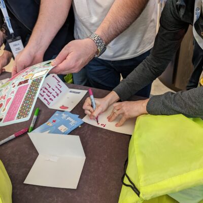 Participants are seen decorating cards with stickers and writing messages for healthcare professionals as part of the Healthcare Heroes scavenger hunt team building event. Bright green donation bags are on the table, ready to be filled with appreciation items.