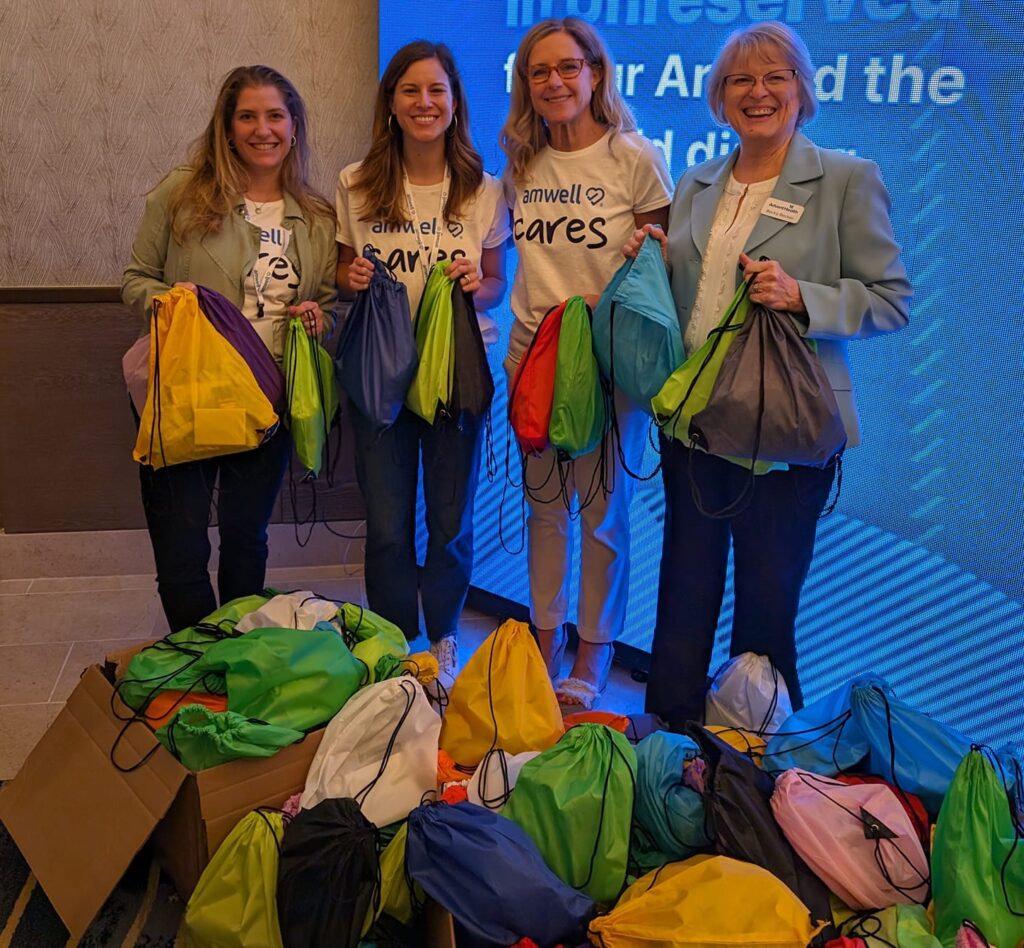 Four participants smiling and holding colorful drawstring bags filled with supplies, standing next to a large pile of completed donation bags as a result of a successful Healtchare Heroes team building scavenger hunt.