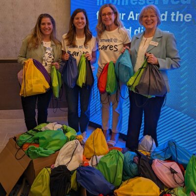 Four participants smiling and holding colorful drawstring bags filled with supplies, standing next to a large pile of completed donation bags as a result of a successful Healtchare Heroes team building scavenger hunt.