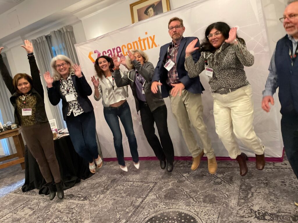 A group of smiling team members enthusiastically jump in the air in front of a CareCentrix banner, celebrating their team building event. They are participating in the Healthcare Heroes program, an interactive team building activity centered on Corporate Social Responsibility, designed to appreciate healthcare workers by assembling donation bags for local hospitals.