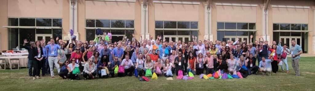 A large group of participants standing outdoors, many holding colorful drawstring bags, gathered in front of a building with tall windows. They are celebrating the accomplishment of a successful Healthcare Heroes team building scavenger hunt.