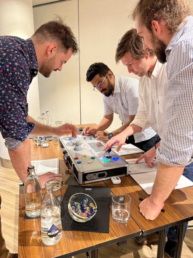 Team members collaborate around a Team Synergy retro gaming console, strategizing and engaging with controls to complete a mini-game challenge during a team building activity.