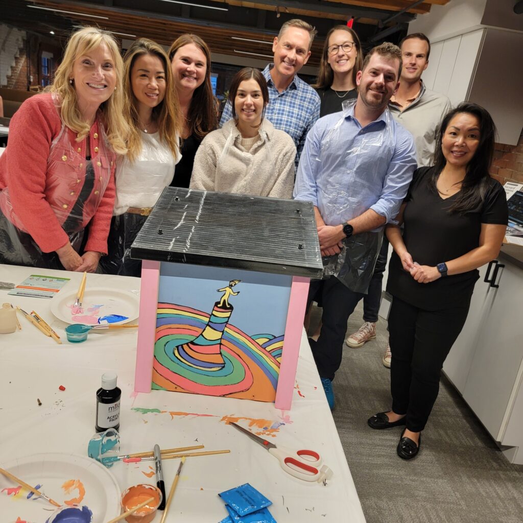 Team proudly poses with their completed Little Free Library during a team building event. The vibrant library features a whimsical design, showcasing teamwork, creativity, and a shared effort to inspire community literacy through the creation of a colorful and engaging book-sharing structure.