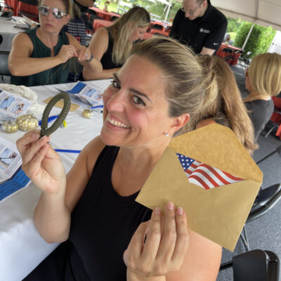 Smiling woman holding an envelope with an American flag and a small parcord bracelet during a corporate team building event, with other participants working in the background.