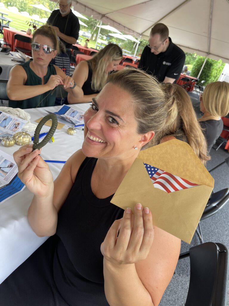 Smiling woman holding an envelope with an American flag and a small parcord bracelet during a corporate team building event, with other participants working in the background.