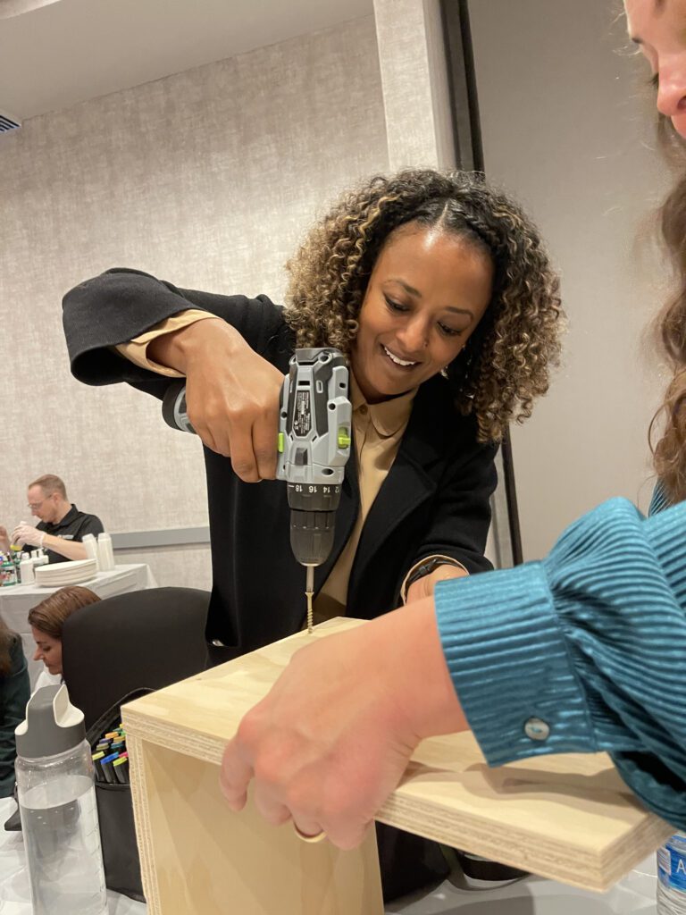 A woman smiles as she uses a power drill to assemble a wooden box, with another person holding the piece steady during the process.