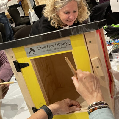 A person paints the outer frame of a wooden Little Free Library box in bright yellow while another person watches. The box is mounted on the table and other materials for decorating the box are scattered in the background.