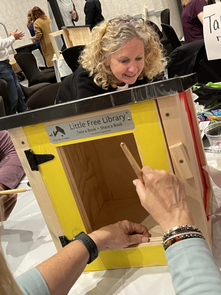 A person paints the outer frame of a wooden Little Free Library box in bright yellow while another person watches. The box is mounted on the table and other materials for decorating the box are scattered in the background.