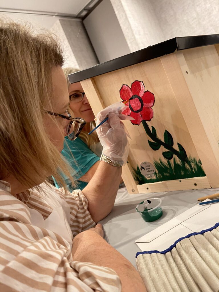 Participant painting a red flower on a wooden Little Free Library as part of the Little Team Library team building event, where teams assemble and decorate book exchange boxes for the community.