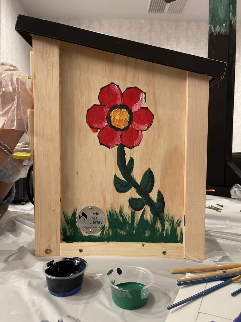 A wooden Little Free Library box in the process of being painted, featuring a bright red flower with a yellow center and green stem on one side. The grass is painted at the base of the box, and small containers of green and black paint sit nearby, along with paintbrushes on the table.