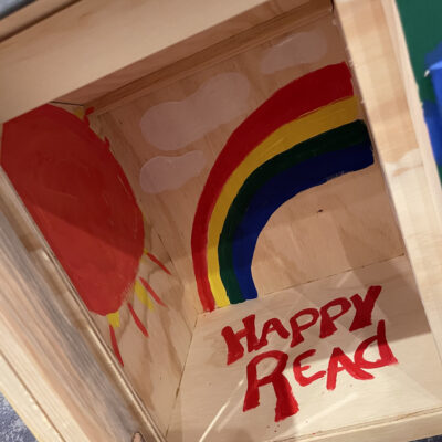 Interior of a decorated Little Free Library featuring a painted rainbow, sun, clouds, and the words 'Happy Read' as part of the Little Team Library team building event.