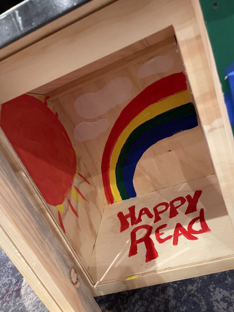 Interior of a decorated Little Free Library featuring a painted rainbow, sun, clouds, and the words 'Happy Read' as part of the Little Team Library team building event.