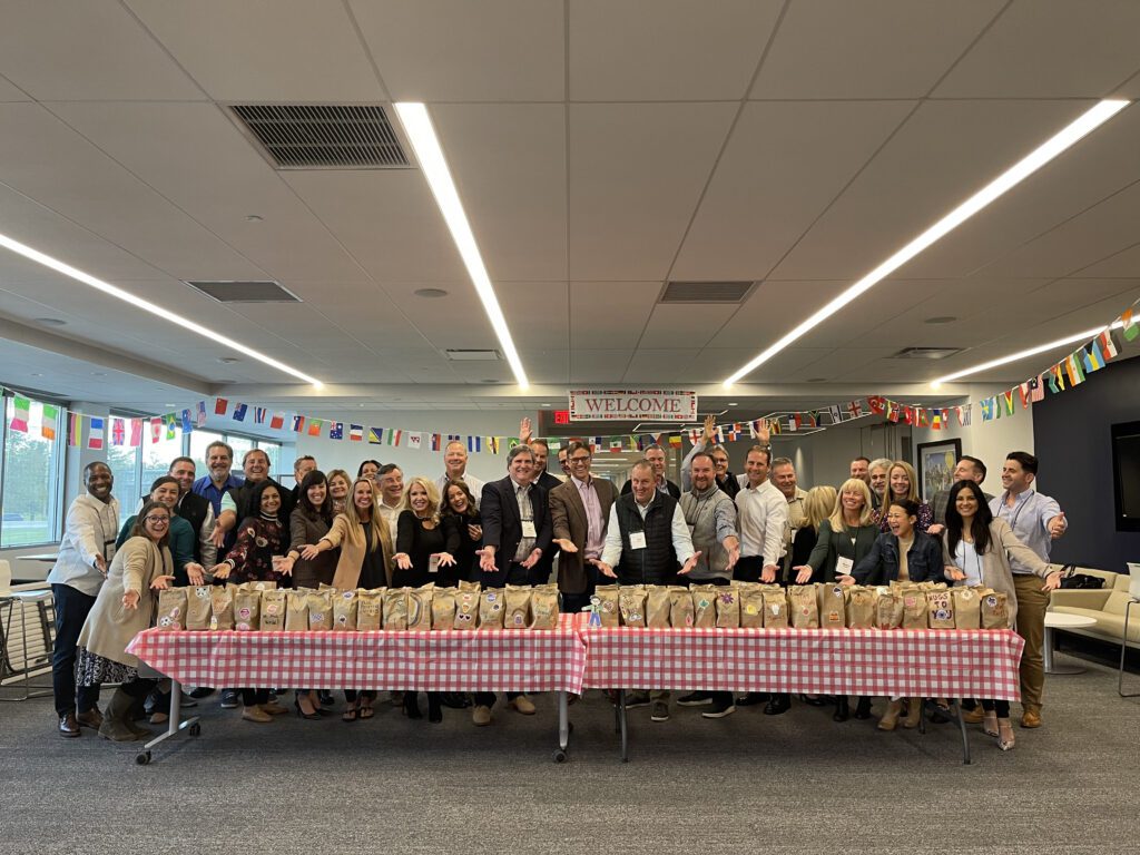 Large group of people posing behind a long table filled with decorated brown paper bags during a corporate giving team building event, with international flags hanging in the background.