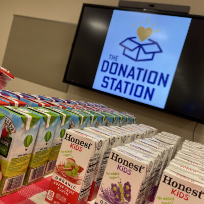 Juice boxes stacked on a table in front of a screen displaying 'The Donation Station' logo during a corporate charitable team building event.