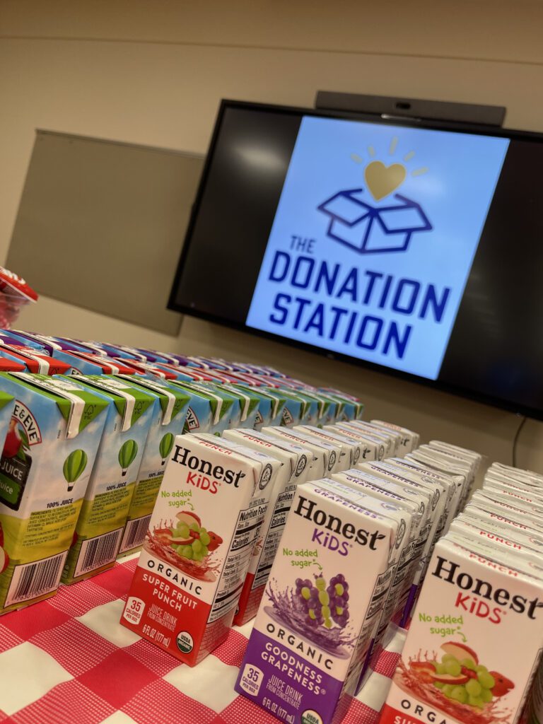 Juice boxes stacked on a table in front of a screen displaying 'The Donation Station' logo during a corporate charitable team building event.