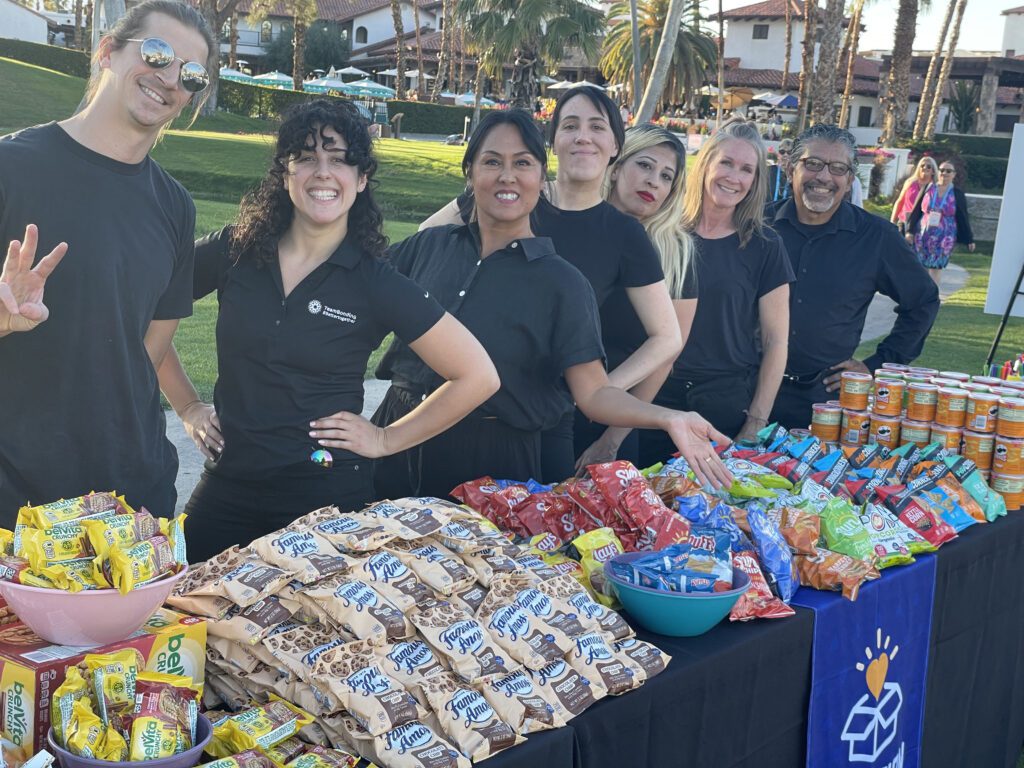 Group of facilitators smiling behind a table filled with snacks and drinks at an outdoor corporate charitable team building event.