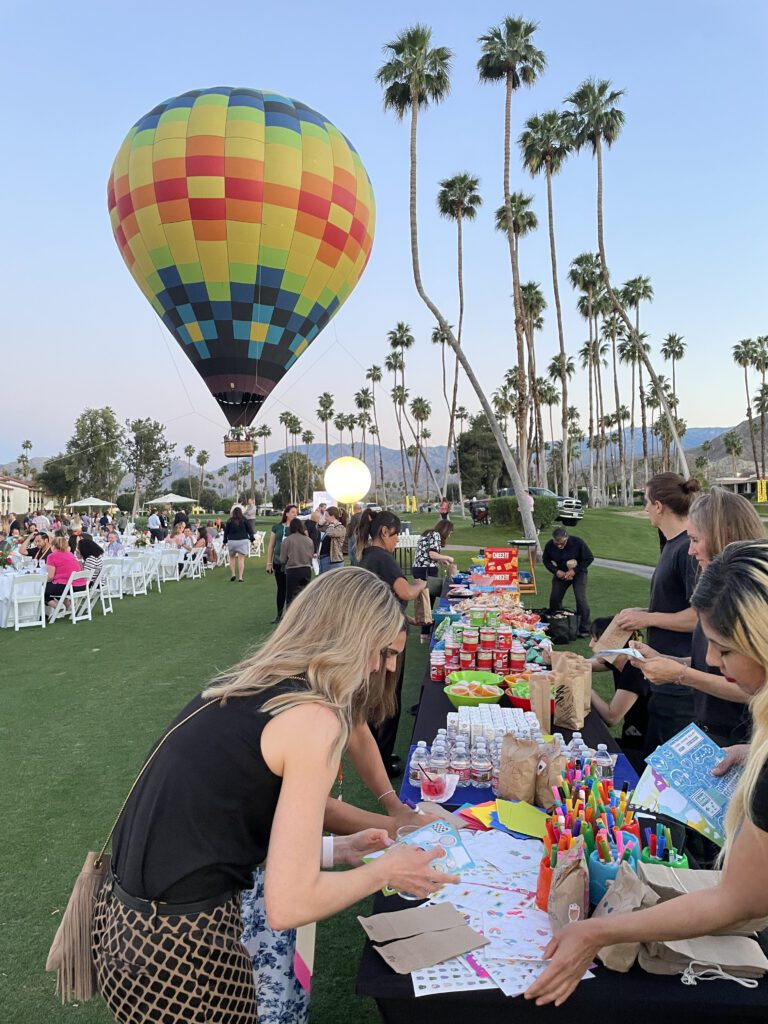 Outdoor corporate charitable team building event with a colorful hot air balloon in the background and participants decorating kits at a table filled with supplies.