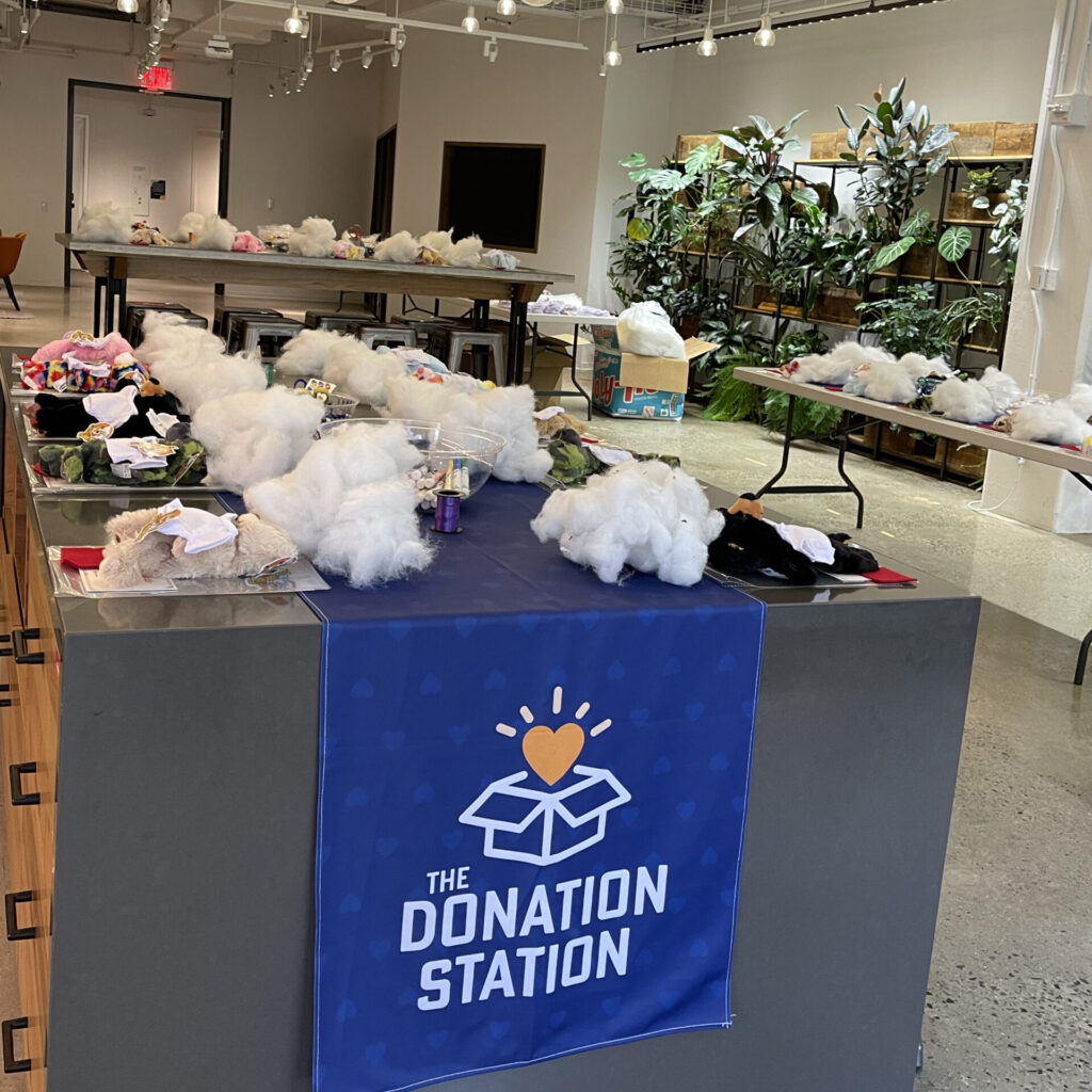 Donation Station setup with tables full of kit materials, including white packing material and small items for assembling donation kits. A banner with 'The Donation Station' is displayed in a brightly lit room, ready for a corporate charitable team building event.