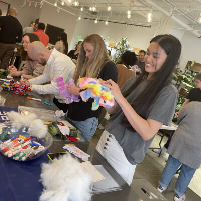 Participants at a Donation Station event assembling colorful donation kits, with supplies like markers and stuffing on the table. A woman in the foreground smiles as she works on a stuffed toy.