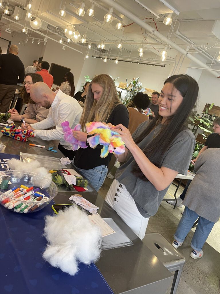 Participants at a Donation Station event assembling colorful donation kits, with supplies like markers and stuffing on the table. A woman in the foreground smiles as she works on a stuffed toy.