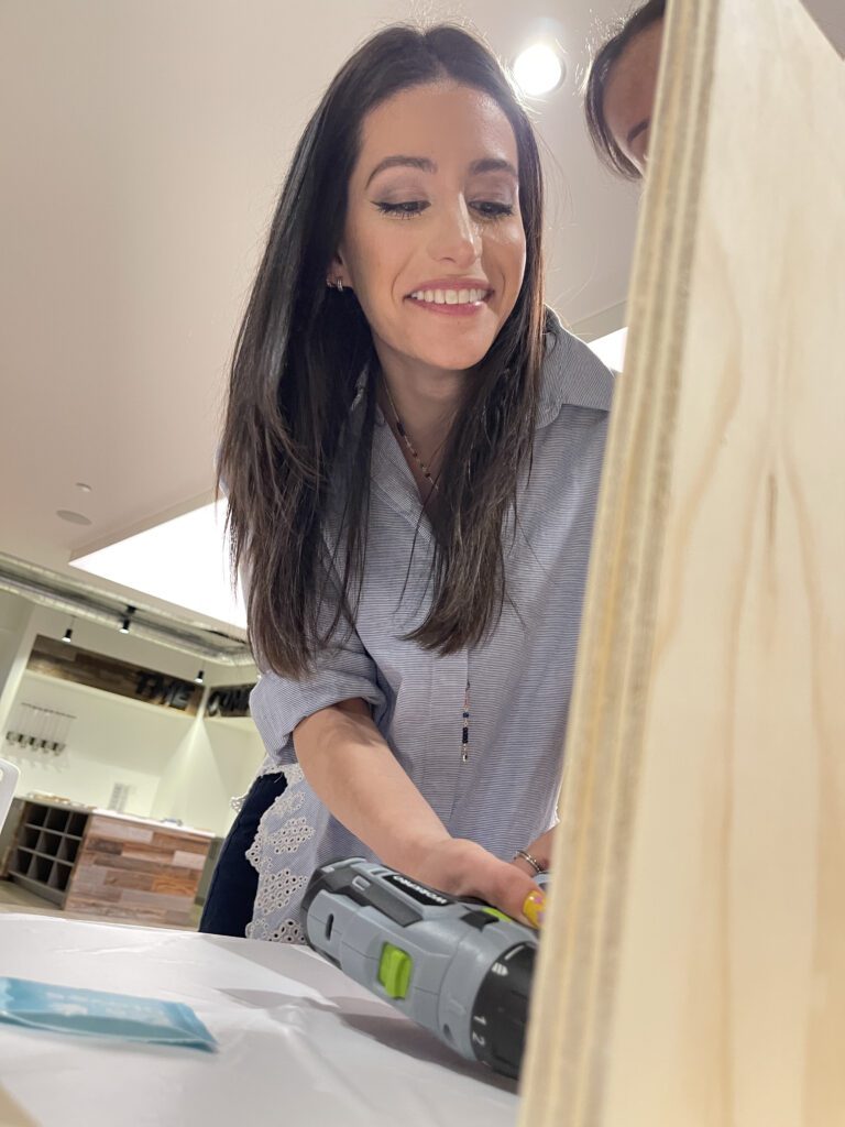 Participant smiling while using a power tool to assemble a Little Free Library during the Little Team Library team building event.