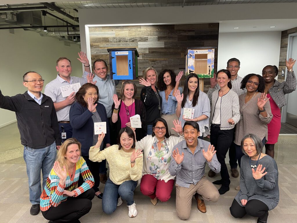 Group of participants smiling and waving in front of two completed Little Free Libraries during the Little Team Library team building event.