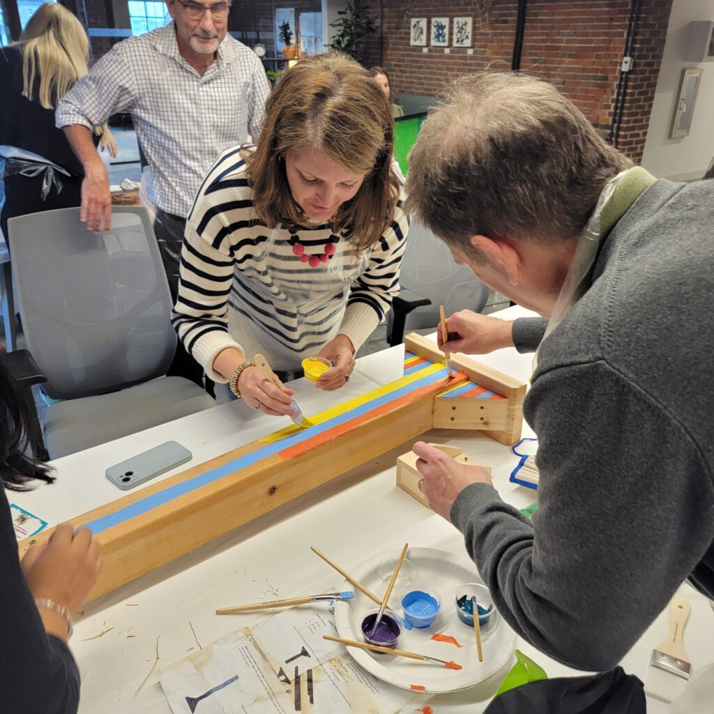 Participants collaborate to paint and decorate a wooden Little Free Library mounting post during a team building event. Each team contributes their creativity to create a cohesive design, fostering teamwork and connection while working toward the shared goal of building a community library.