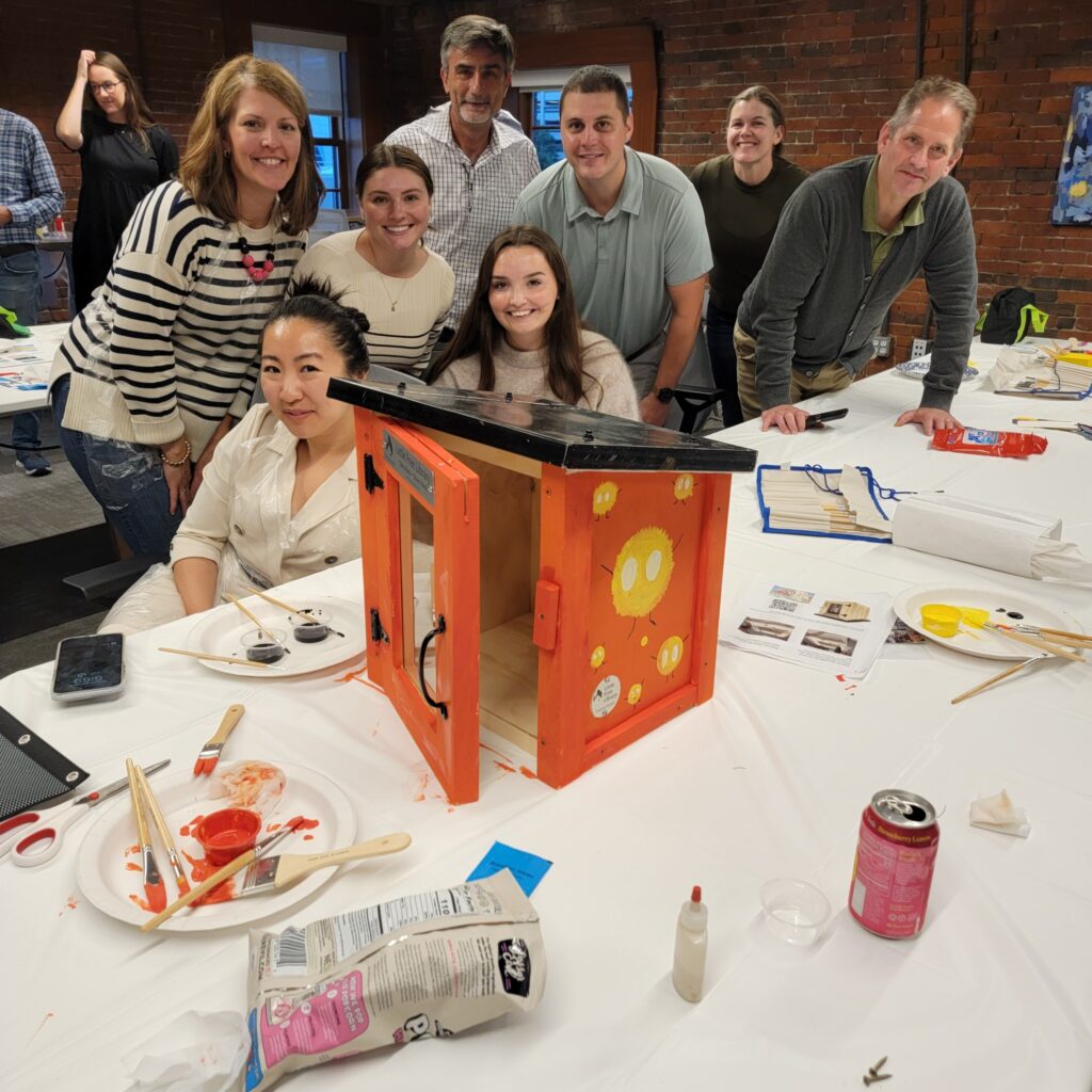 Participants gather around a table to mix and paint with vibrant colors during a creative team building activity. The hands-on session encourages collaboration and artistic expression as teams work together to design elements for a Little Free Library, promoting teamwork and community engagement.