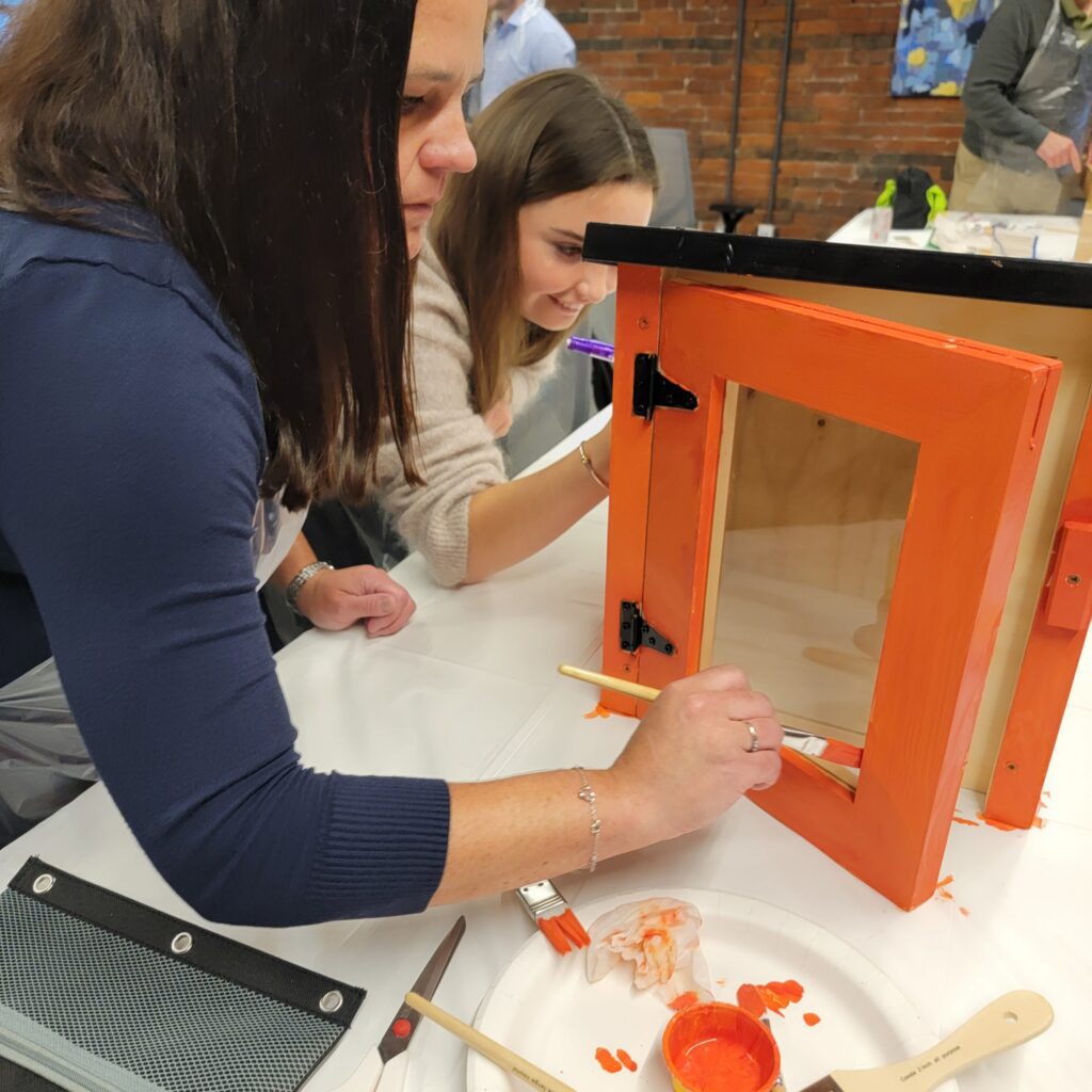 Participants focus on painting a Little Free Library door with bright orange during a collaborative team building event. The hands-on activity fosters creativity and teamwork as teams work together to build and decorate the library for community use.