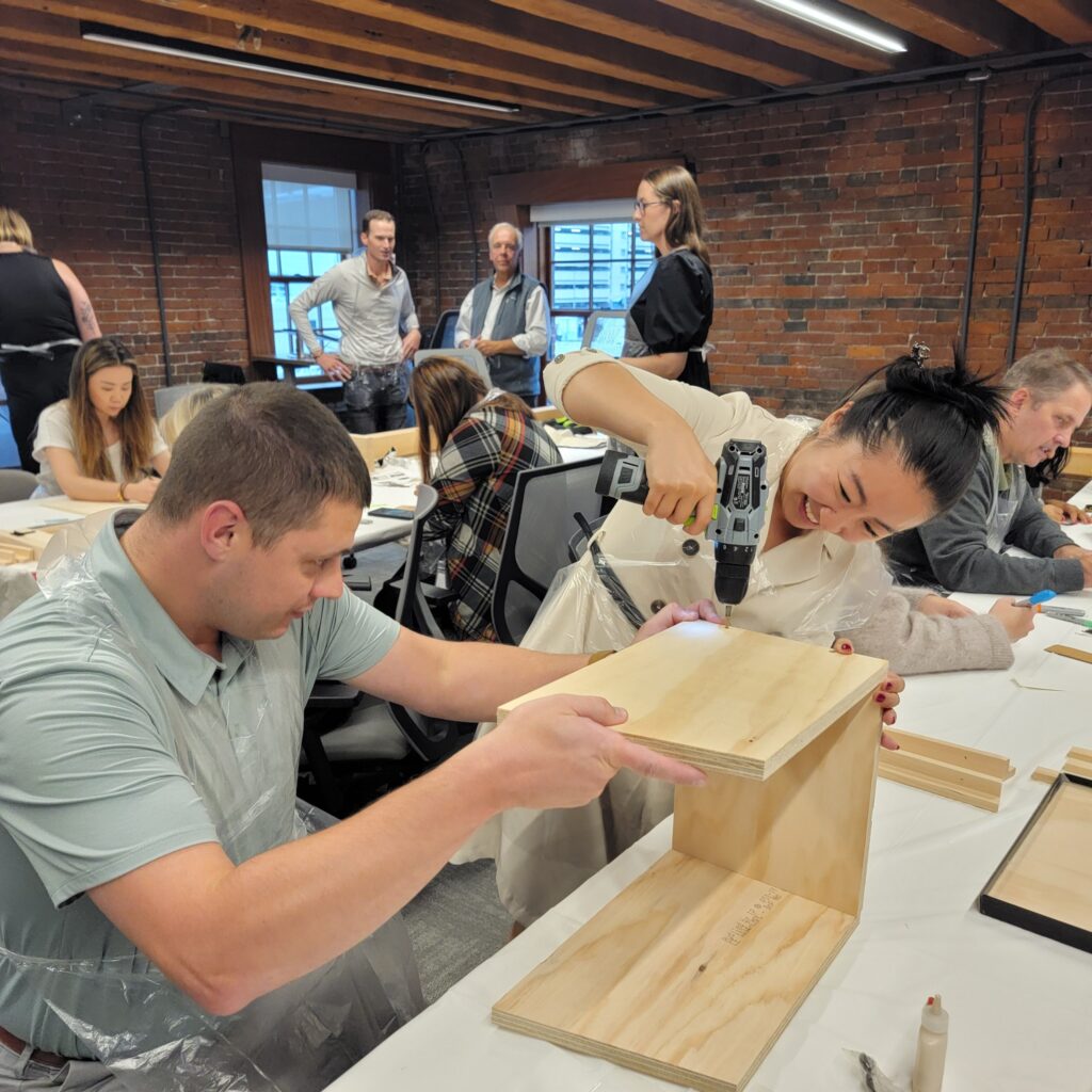 Enthusiastic participants work together to assemble a Little Free Library during a team building event. One team member uses a power drill while another holds the wooden pieces steady, demonstrating teamwork, coordination, and problem-solving as they build the library structure.