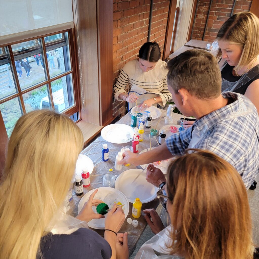 Participants gather around a table to mix and paint with vibrant colors during a creative team building activity. The hands-on session encourages collaboration and artistic expression as teams work together to design elements for a Little Free Library, promoting teamwork and community engagement.