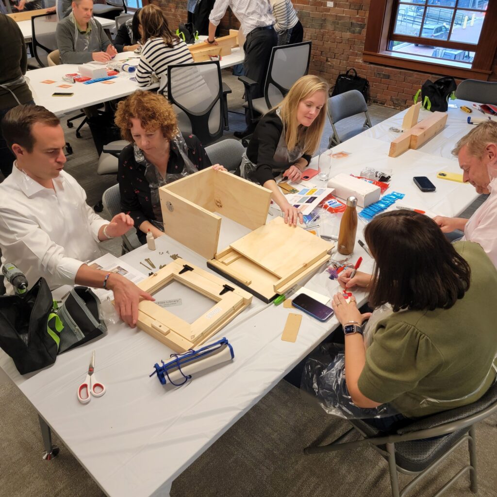 Teams collaborate to assemble a Little Free Library during a hands-on team building event. Participants work together to construct the wooden structure, following instructions and dividing tasks to create a functional and beautifully decorated library for the community.